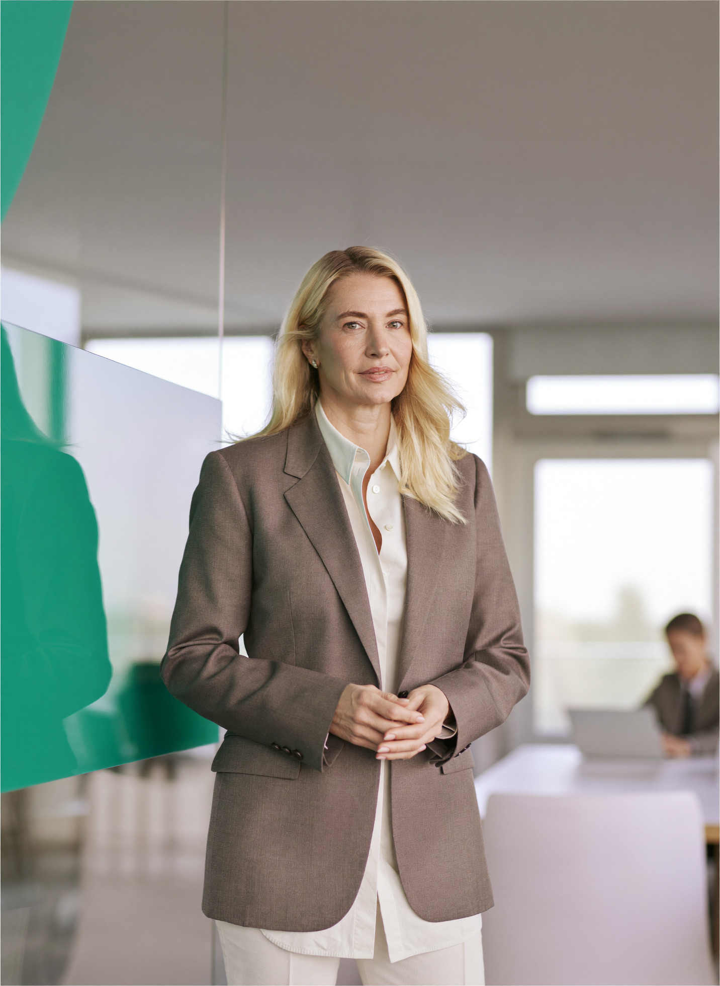A senior compliance officer stands in a bright bank office beside a teal glass panel, a colleague at a desk behind her.