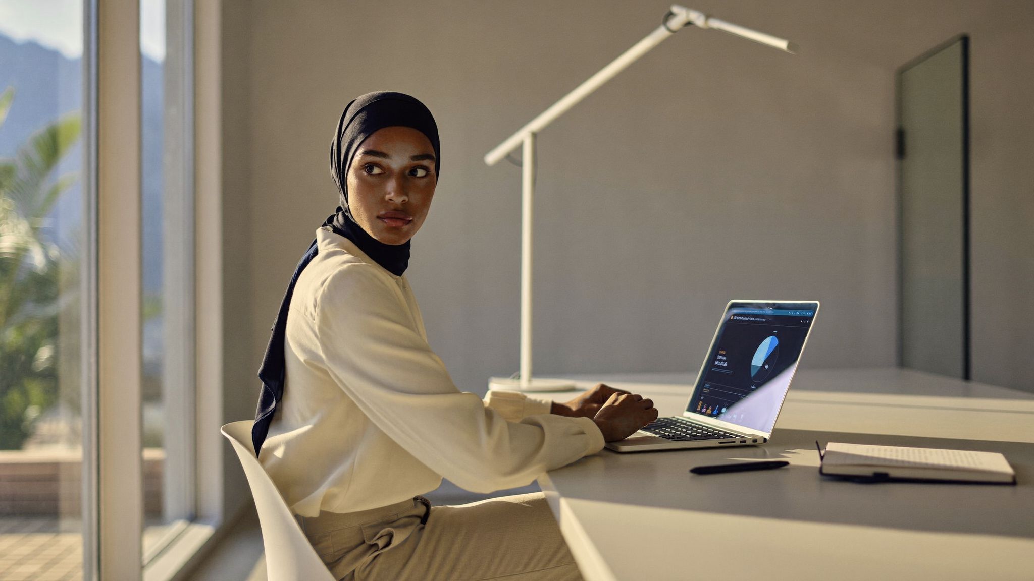 مسؤولة تعلّم على مكتب تغمره الشمس، ترفع نظرها عن حاسوب محمول تظهر عليه لوحة متابعة.