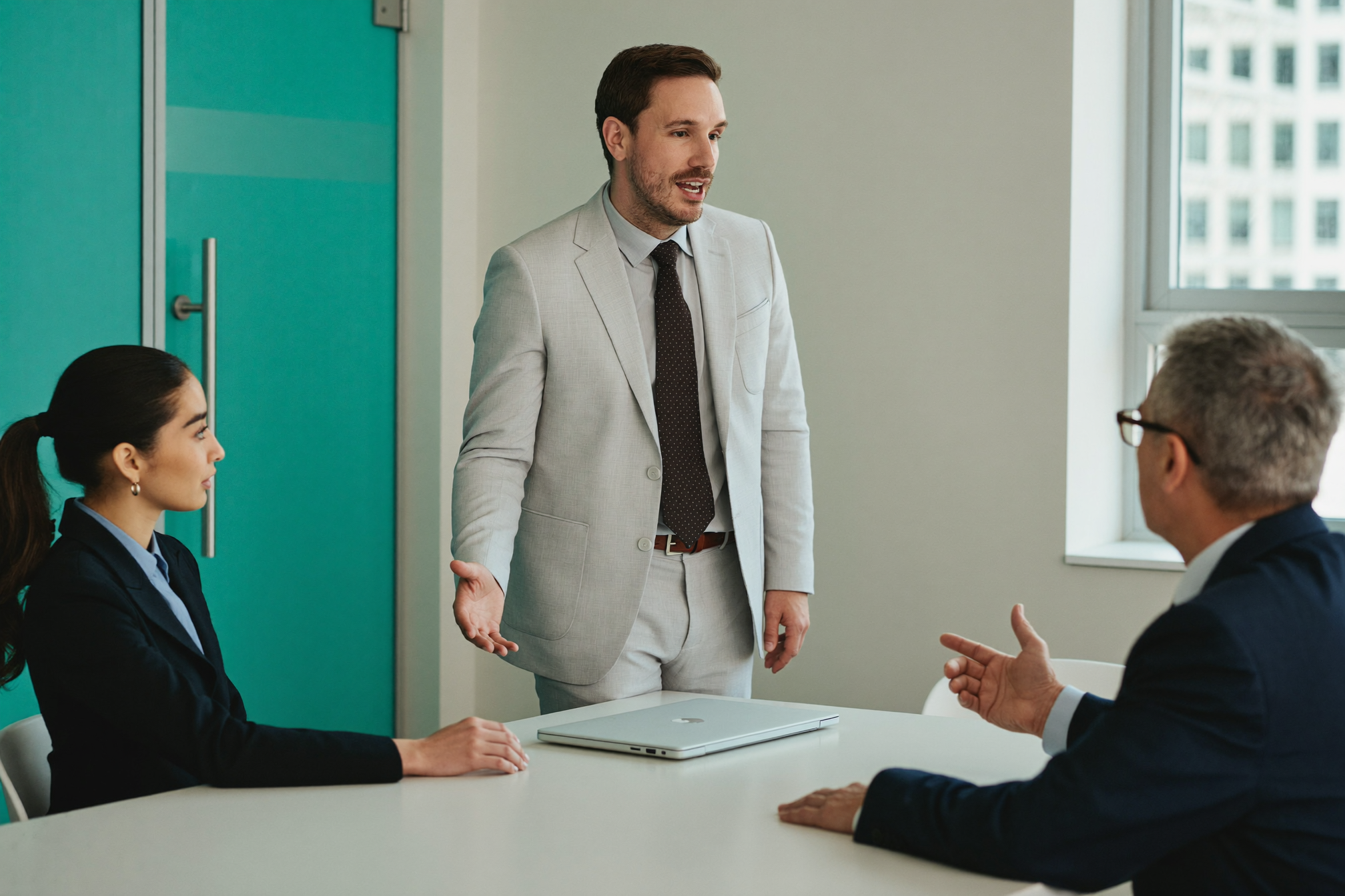 Three colleagues confer around a bright conference table, one standing as he explains and two seated responding across the table.